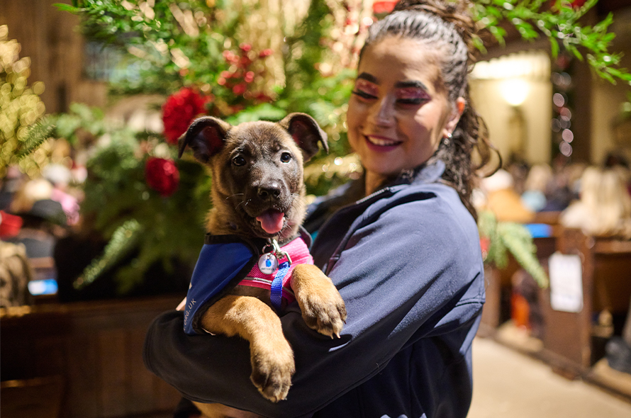 A woman joyfully holds a puppy at a festive Christmas party, surrounded by holiday decorations and cheerful guests.